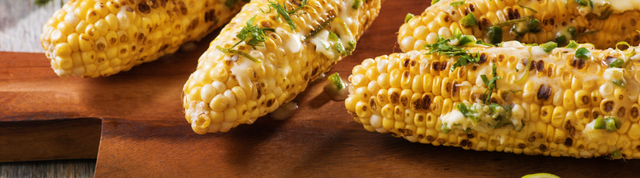four grilled corn on the cob with melted butter and herbs on a wooden plate