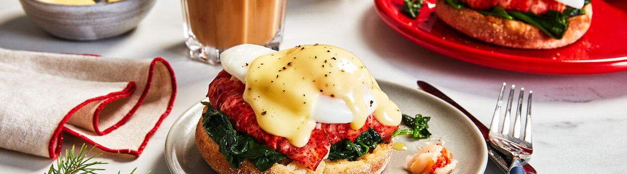 Marble surface with two red plates topped with classic Eggs Benedict, with a layer of lobster beneath the poached egg, and two lattes in the background.