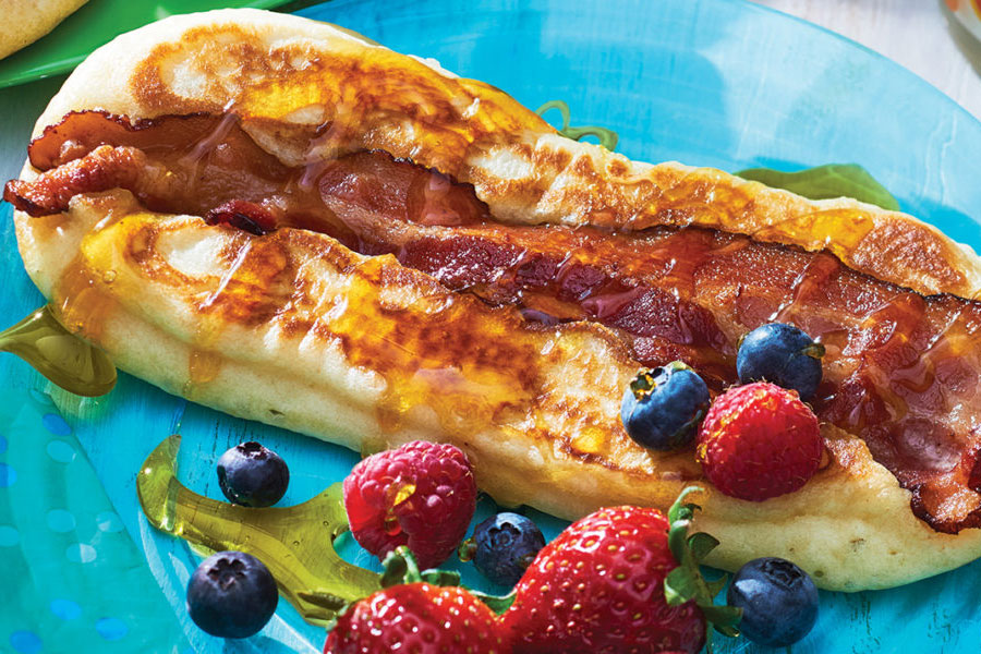 Blue plate of pancakes with bacon slice baked in, covered in syrup and berries.