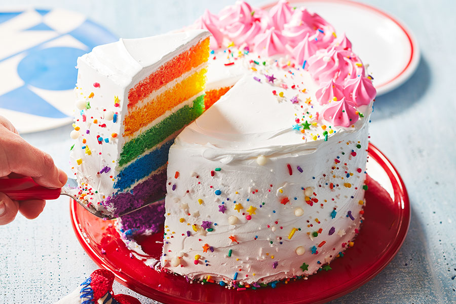 5-layer rainbow cake with rainbow sprinkles and pink rosettes on top sitting on red plate with slice being taken out