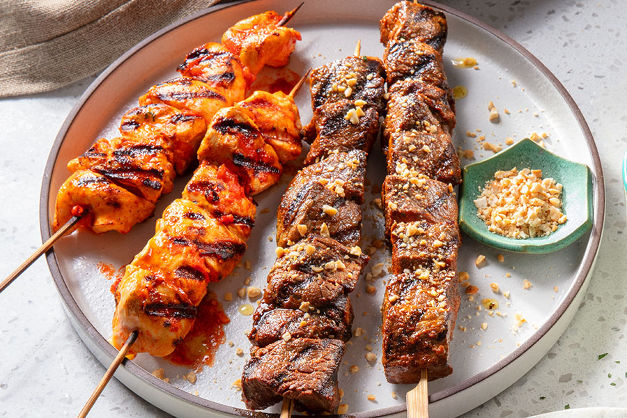 Three Caribbean-inspired kabobs including pork and chicken with glazes and pineapple pieces on cream coloured platters with a napkin off to the side.