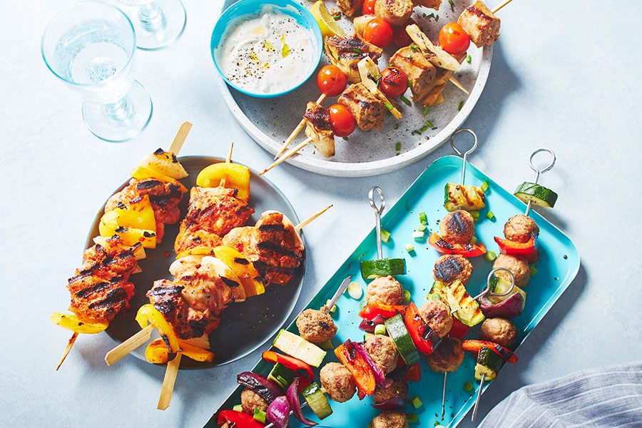 Three kinds of European-inspired kabobs on three different platters: one thatâ€™s gray, blue and white with water glasses in the background and a blue napkin.
