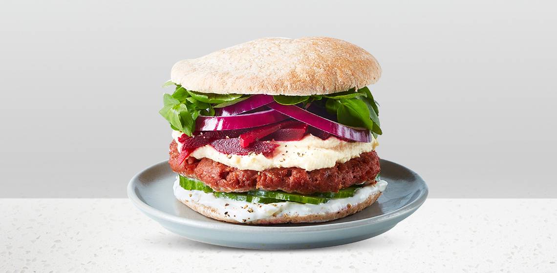 Falafel-Style Burger: soft whole wheat pita or seeded kaiser roll stacked with a Beyond Meat Plant-Based Burger Patty, hummus, pickled beets, cucumber slices, tahini sauce or tzatziki, red onion, and arugula.