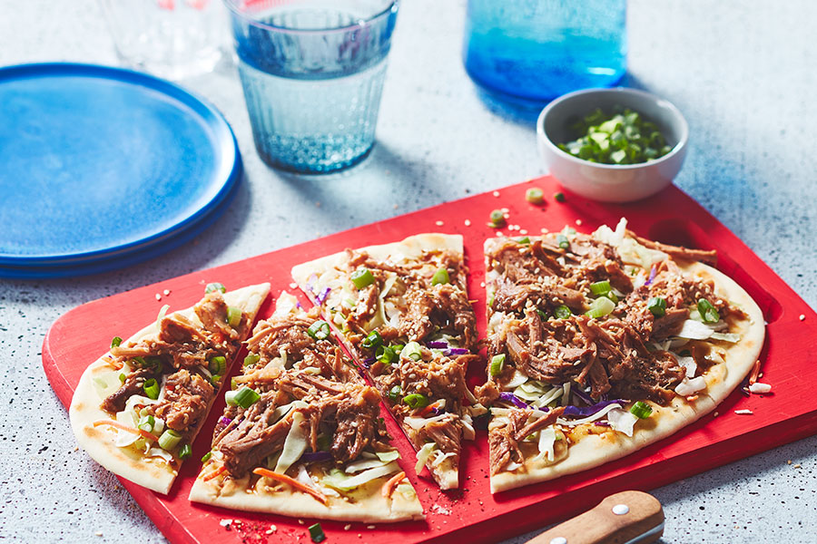 Korean-style, pulled pork topped flatbread garnished with coleslaw, green onions and sesame seeds sitting on a red cutting board with a knife next to it.