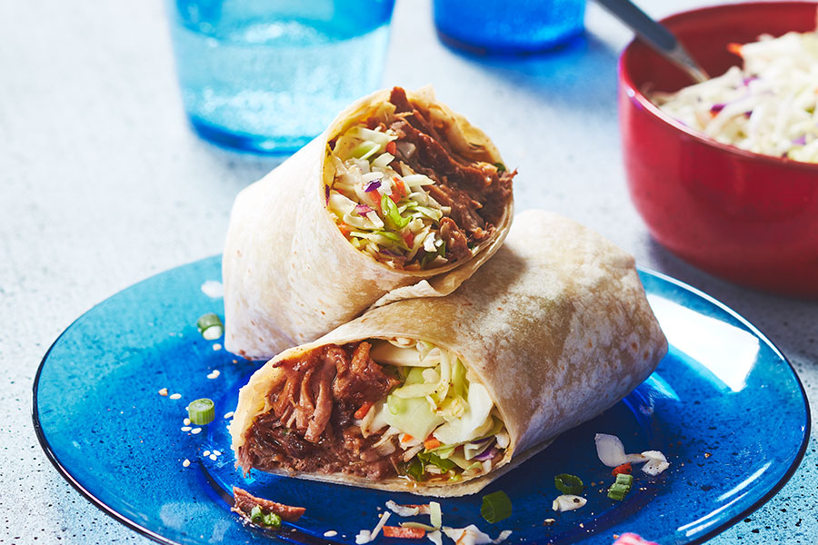 Two halves of a wrap style sandwich filled with Korean-style marinated pulled pork, coleslaw, green onions, and sesame seeds sitting on a clear blue dinner plate.