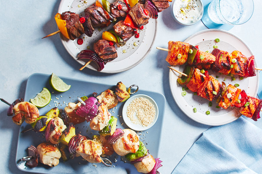 Three North-American-flavoured skewers including one beef, one chicken and one salmon on blue and white and gray plates with water glasses off to the side.