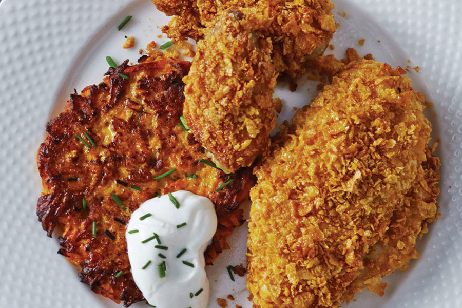 Cornflake crumb coated oven fried chicken on a white plate sitting next to a sweet potato and carrot rosti.
