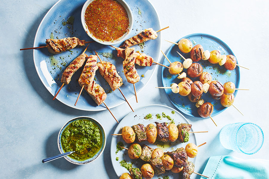 The South American-inspired kabobs, one with beef and potato, one potato and garlic, the last pork on blue plates with a bowl of chimichurri to the side.