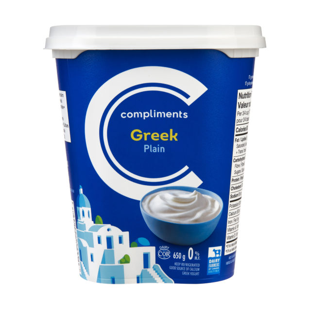 A white tub of Compliments Plain 0% Greek Yogurt with an illustration of a Greek village and whitewashed houses by the sea.