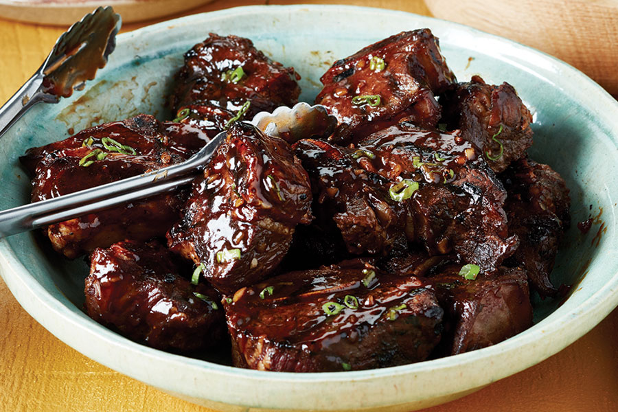 A light-blue bowl full of glazed Asian-style BBQ beef short ribs with tongs for serving. 