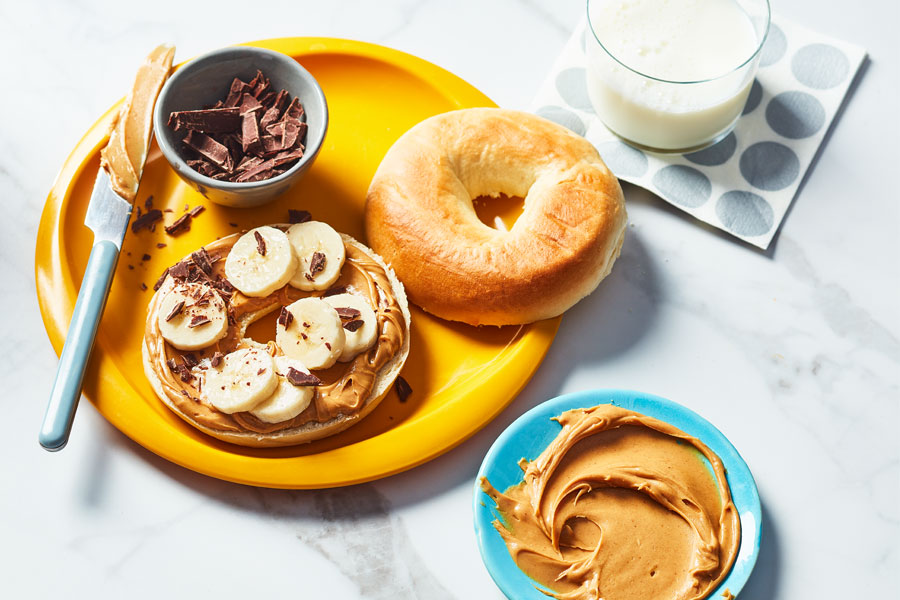 Open faced bagel covered with peanut butter, sliced bananas and chocolate on a yellow plate.