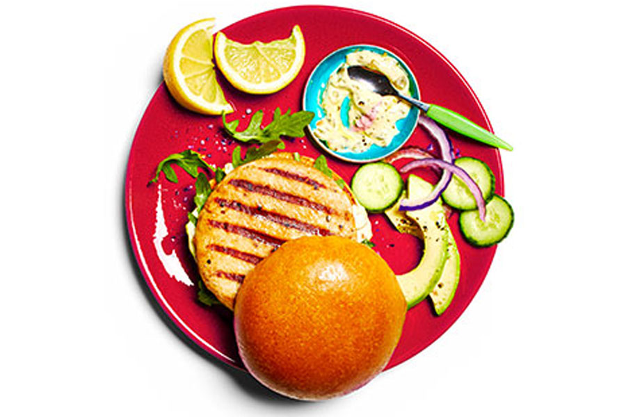 Cooked and dressed wild sockeye salmon burger on a red plate next to sliced avocado and onion.