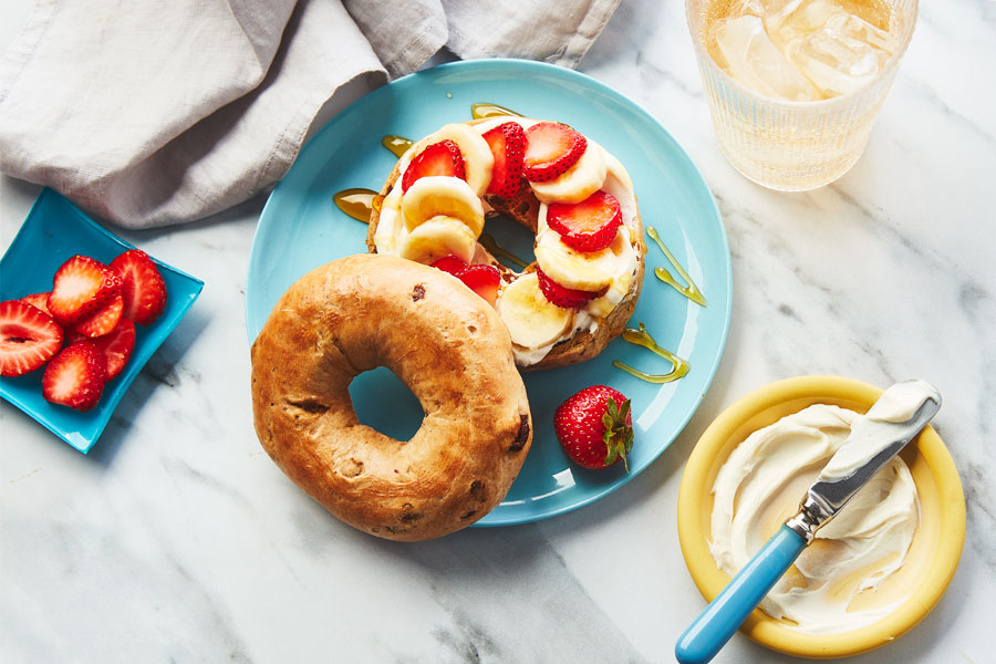 Cinnamon swirled bagel topped with Greek yogurt, cream cheese, strawberry and banana slices and a drizzle of honey on a pale blue plate.