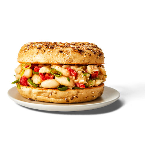 Multigrain bagel topped with canned white beans, roasted red peppers, green onions, minced garlic and olive oil on a white plate.