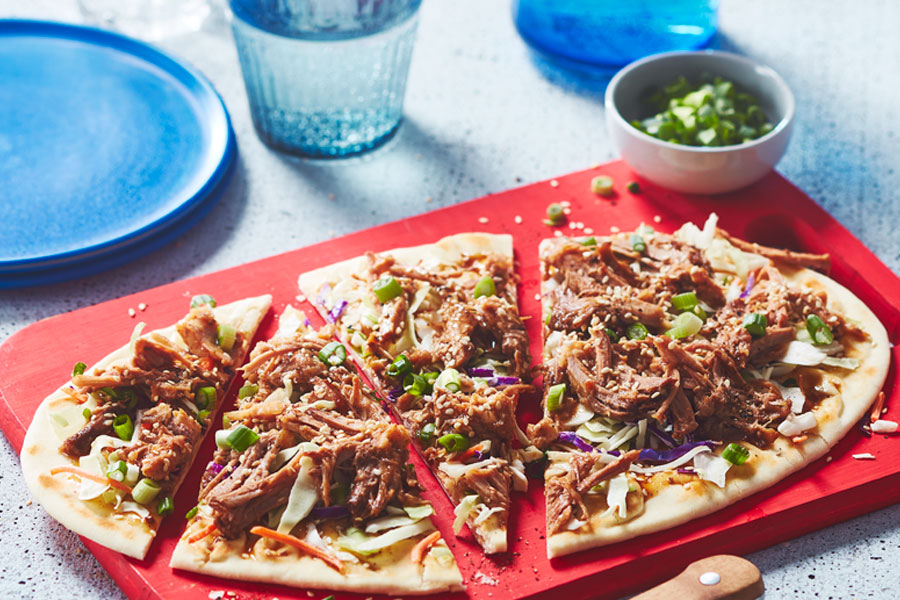 Korean-style pulled pork–topped flatbread, garnished with coleslaw, green onions, and sesame seeds, sitting on a red cutting board with a knife next to it.