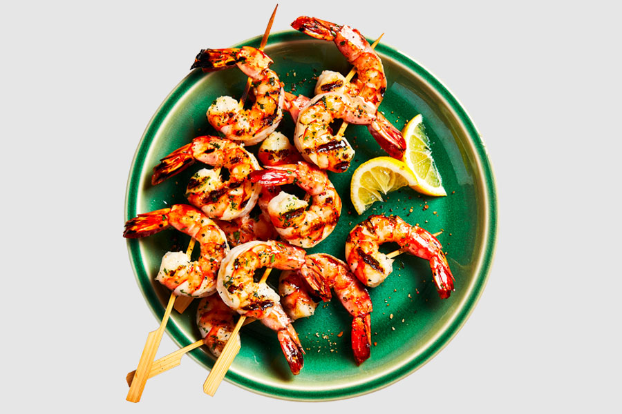 A round green plate with three cooked shrimp skewers laid on top, and cut limes for garnish.