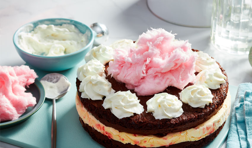 Marble tabletop with a blue cutting board topped with a whole chocolate cake, a middle layer of ice cream and a whipped cream and cotton-candy decorated top.