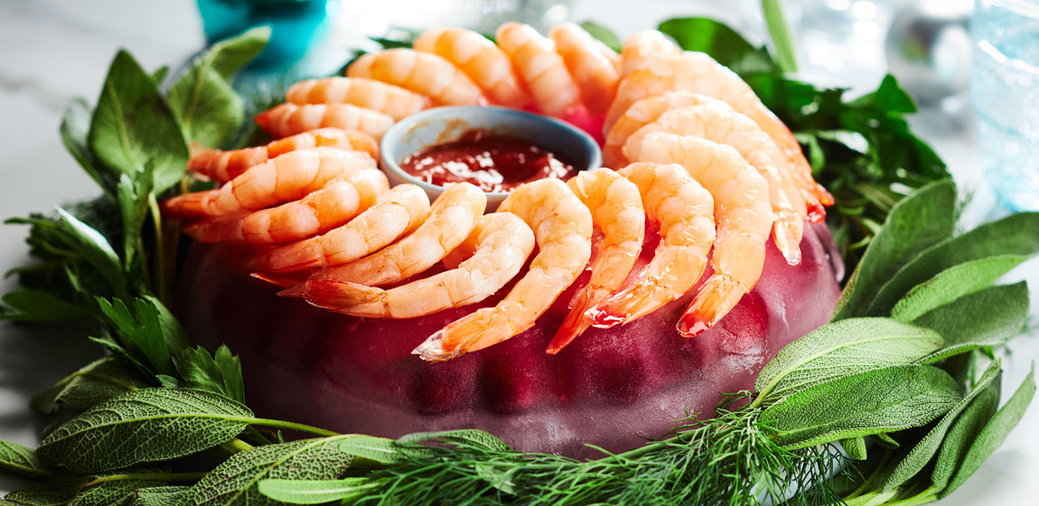 Platter with a round ice ring with frozen cranberries inside, edged with fresh green herbs and topped with Compliments Naturally Simple Cooked Pacific White Shrimps.