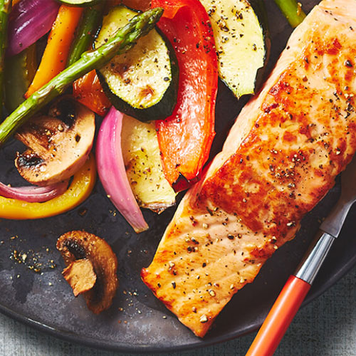 A marinated salmon fillet on a grey plate next to a side of vegetables flecked with fresh pepper.