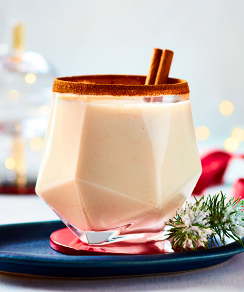 Angular gem-cut drinking glass with a cinnamon rim, milky-coloured whiskey drink and cinnamon stick garnish on a set holiday table.