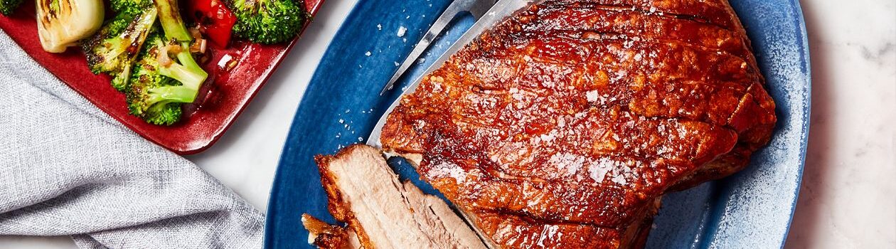 Slices of crispy pork belly on a gray platter with a bowl of charred bok choy and broccoli beside it in a red serving platter on a white countertop.