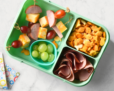 Green bento box filled with skewers of prepared sausage, cheese and grapes, mini cheese crackers and smoked ham slices.