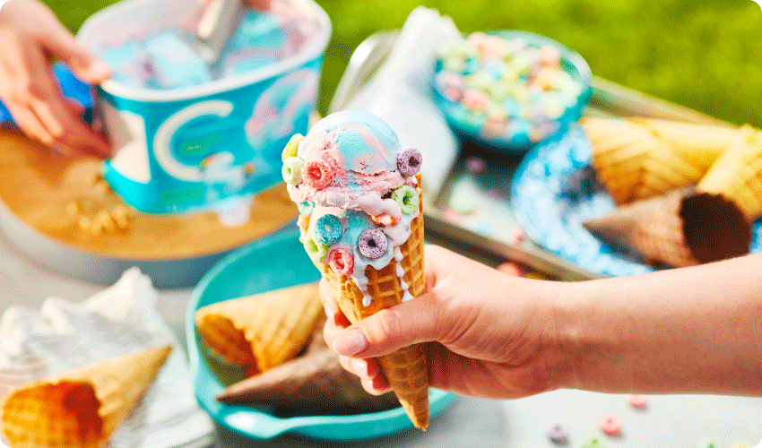 young hand holding waffle ice cream cone with unicorn twirl ice cream studded with fruity hoops cereal around the edges.