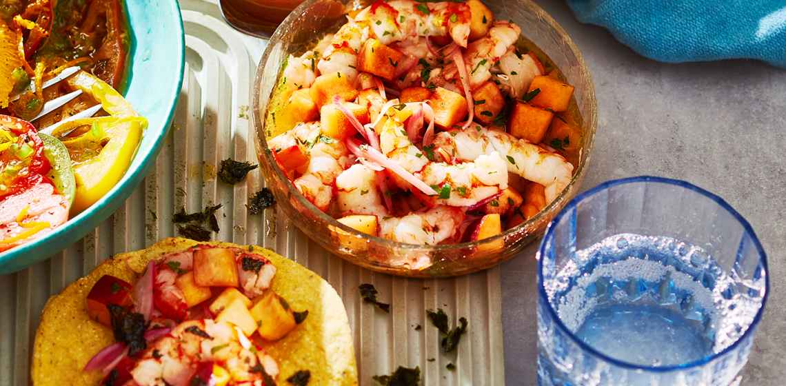 Clear bowl full of shortcut grilled shrimp and peach ceviche next to a toasted, topped tortilla.