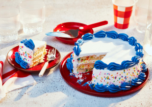 Triple-layer confetti cake iced in white and pink with sprinkles, sitting atop a round red dessert platter with a slice cut out on a smaller plate with a fork next to it.