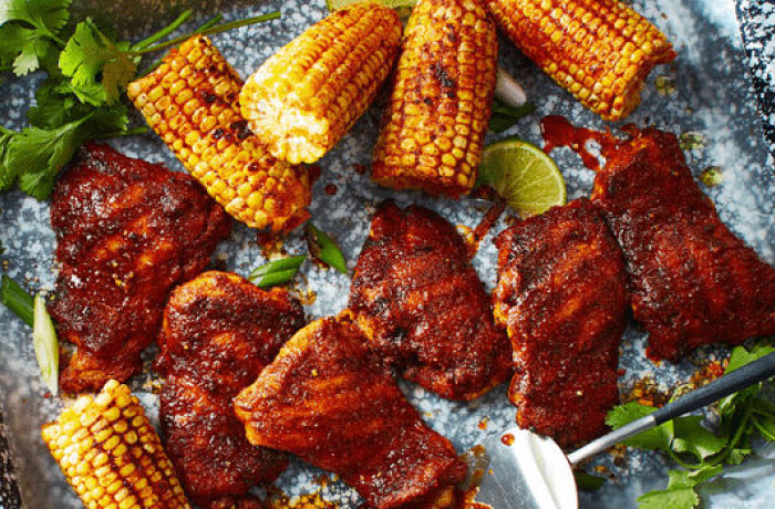 Paprika grilled chicken thighs with corn on the cob on a speckled baking sheet.