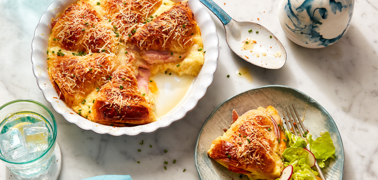 Ham and cheese brunch or breakfast croissant strata in a white baking dish with a serving next to it on a gray dish with a side salad.