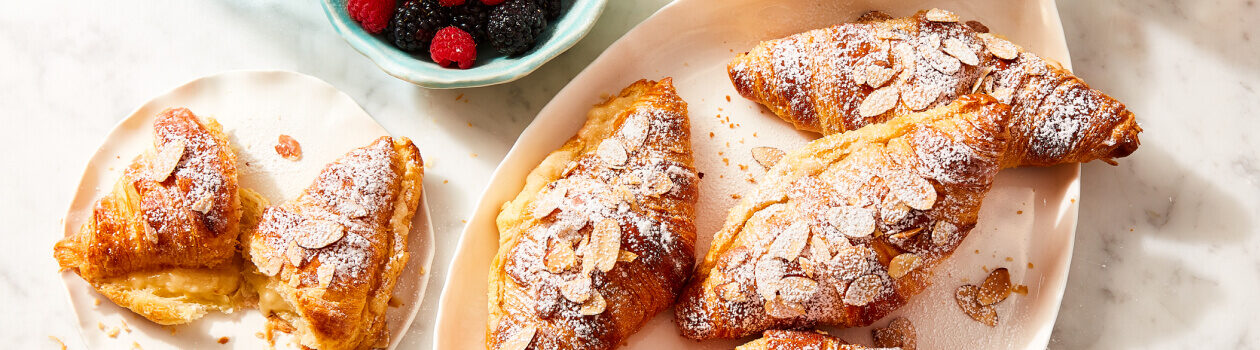 Speedy almond croissants topped with slivered almond slices in a cream serving dish next to a plate with one croissant on it.