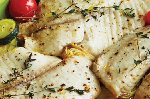 White fish fillets in a green baking dish with cherry tomatoes, herbs, zucchini and artichoke hearts.