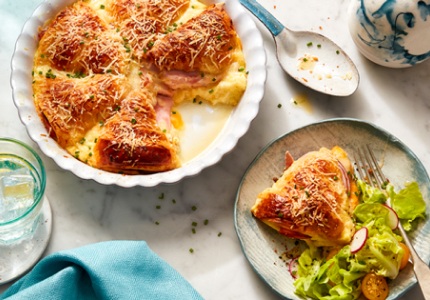 A white dish with Double Ham and Cheese Croissant Strata with a serving scooped out on a small white side plate with a side salad.